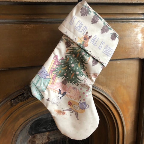 A cream coloured Christmas stocking hung from a mantlepiece. on the stocking is a festive, hand drawn image of 4 playful bunnies causing mischief around a Christmas tree, under the phrase 'all is calm, all is bright'.