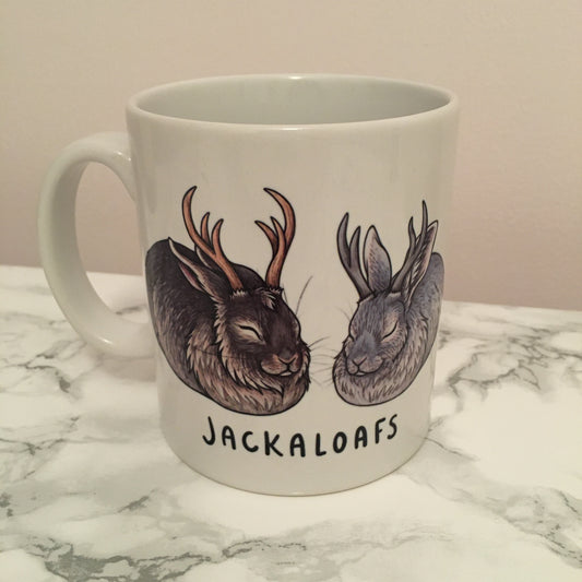 A white mug with two jackalope rabbits in the laof position. One is brown and the other is grey. Written below the jackalopes is JACKALOAFS in black