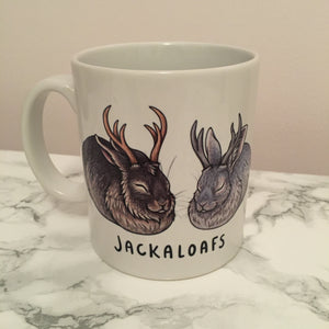 A white mug with two jackalope rabbits in the laof position. One is brown and the other is grey. Written below the jackalopes is JACKALOAFS in black
